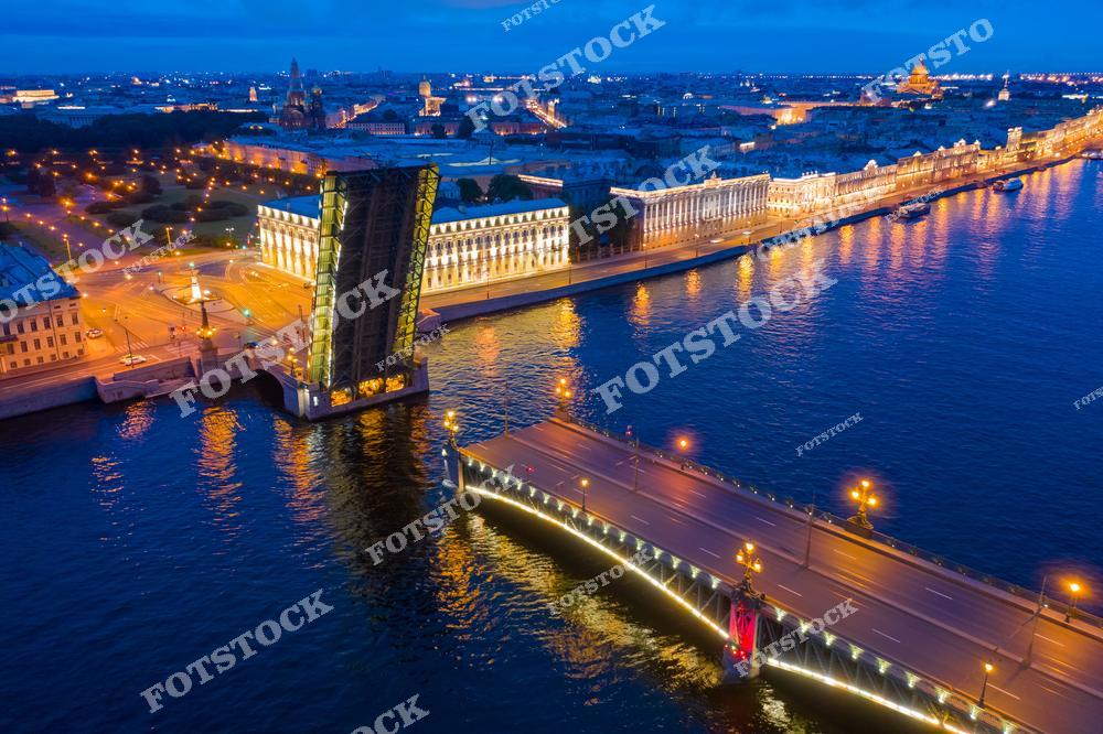 На фотографии изображен разведённый Троицкий мост, соединяющий центр Санкт-Петербурга с Петроградской стороной. Этот мост, украшенный декоративной подсветкой, является одним из старейших и самых красивых мостов города. На заднем плане видны исторические здания, такие как Зимний дворец и Исаакиевский собор, освещенные вечерними огнями. Отражения в воде добавляют фотографии магии и романтики.